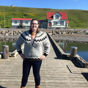One of the Snorri alumni interns posing on the dock in the Hofsós harbour, in Iceland, in front of one of the museum buildings. It is a sunny day with blue skies. The intern is wearing one of the infamous Icelandic wool sweaters, the lopapeysa.